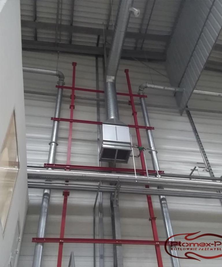 Nagrzewnica powietrza zamontowana na konstrukcji nośnej w hali przemysłowej z widocznymi rurami instalacyjnymi.