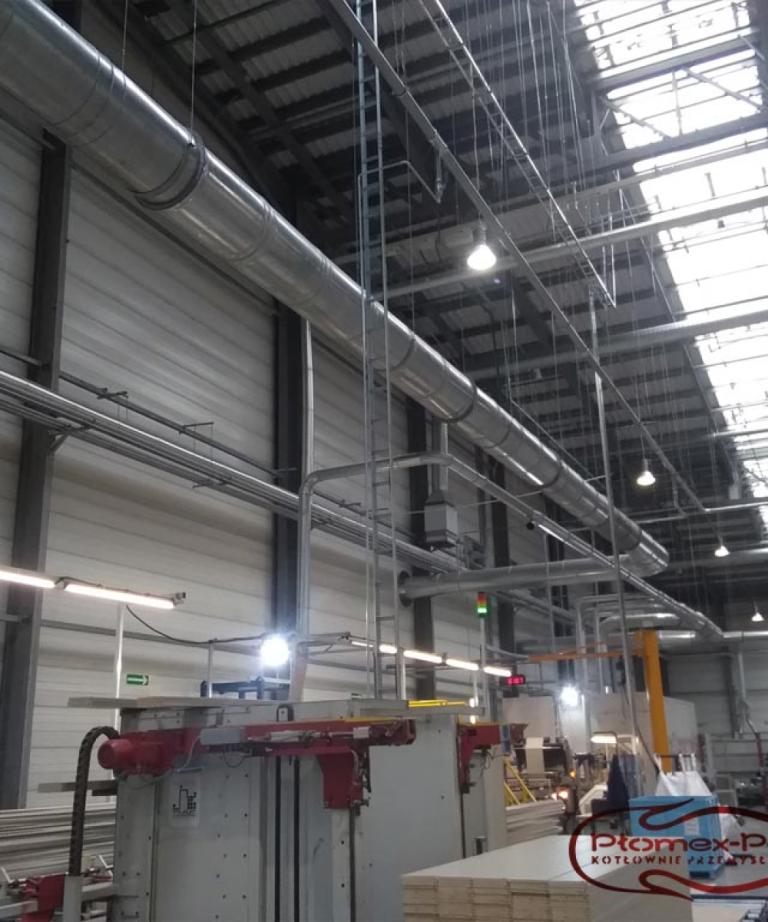 Szeroki widok instalacji rur doprowadzających ciepłe powietrze do maszyn wewnątrz hali produkcyjnej.