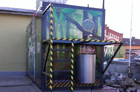 Nowoczesna kontenerowa kotłownia na pellet firmy Plomex-Pol z grafiką lasu na obudowie, zainstalowana na zewnątrz budynku.