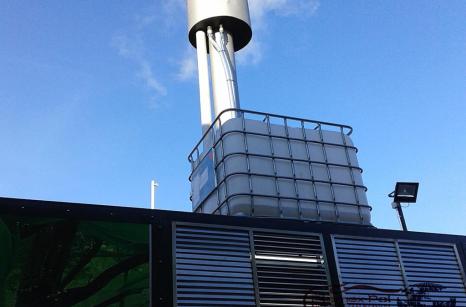 Zewnętrzny element kontenerowej kotłowni na pellet w Pile z widocznym zbiornikiem na glikol typu paletopojemnik IBC oraz stalowym kominem na tle nieba.