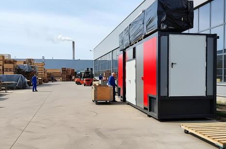 Przemysłowa kotłownia kontenerowa o mocy MAC-720kW z czerwono-szarymi panelami, gotowa do transportu, stojąca na placu fabrycznym.