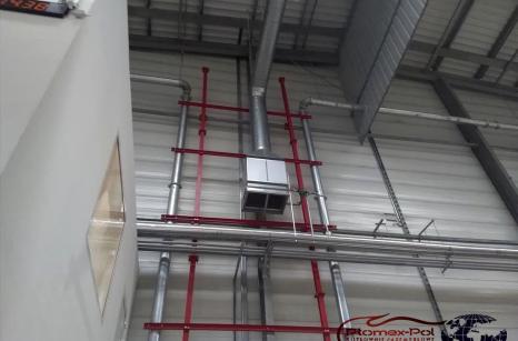 Nagrzewnica powietrza zamontowana na konstrukcji nośnej w hali przemysłowej z widocznymi rurami instalacyjnymi.
